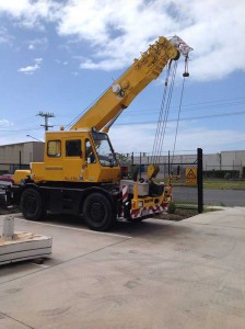 Yellow mobile crane truck with extended boom parked at construction site against cloudy blue sky.